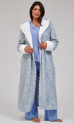 A woman stands barefoot against a light gray studio background wearing a long textured light blue robe with white faux-fur trim over a light blue pajama set.