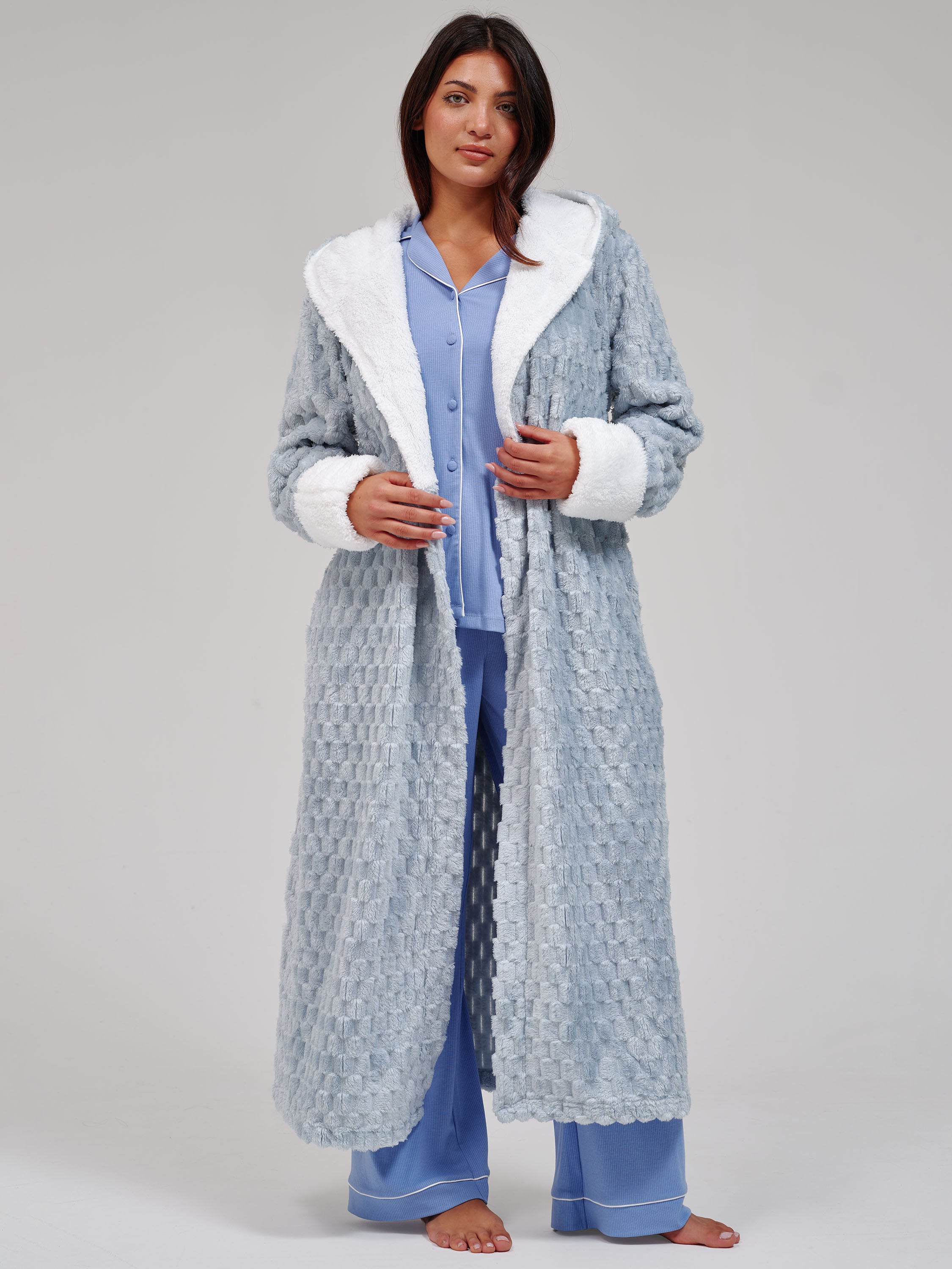 A woman stands barefoot against a light gray studio background wearing a long textured light blue robe with white faux-fur trim over a light blue pajama set.