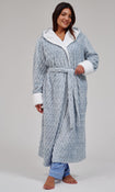 A woman modeling a light blue textured waffle-weave hooded bathrobe with white faux-fur trim on the hood, cuffs, and waist tie, paired with blue pajama pants against a neutral gray studio background.