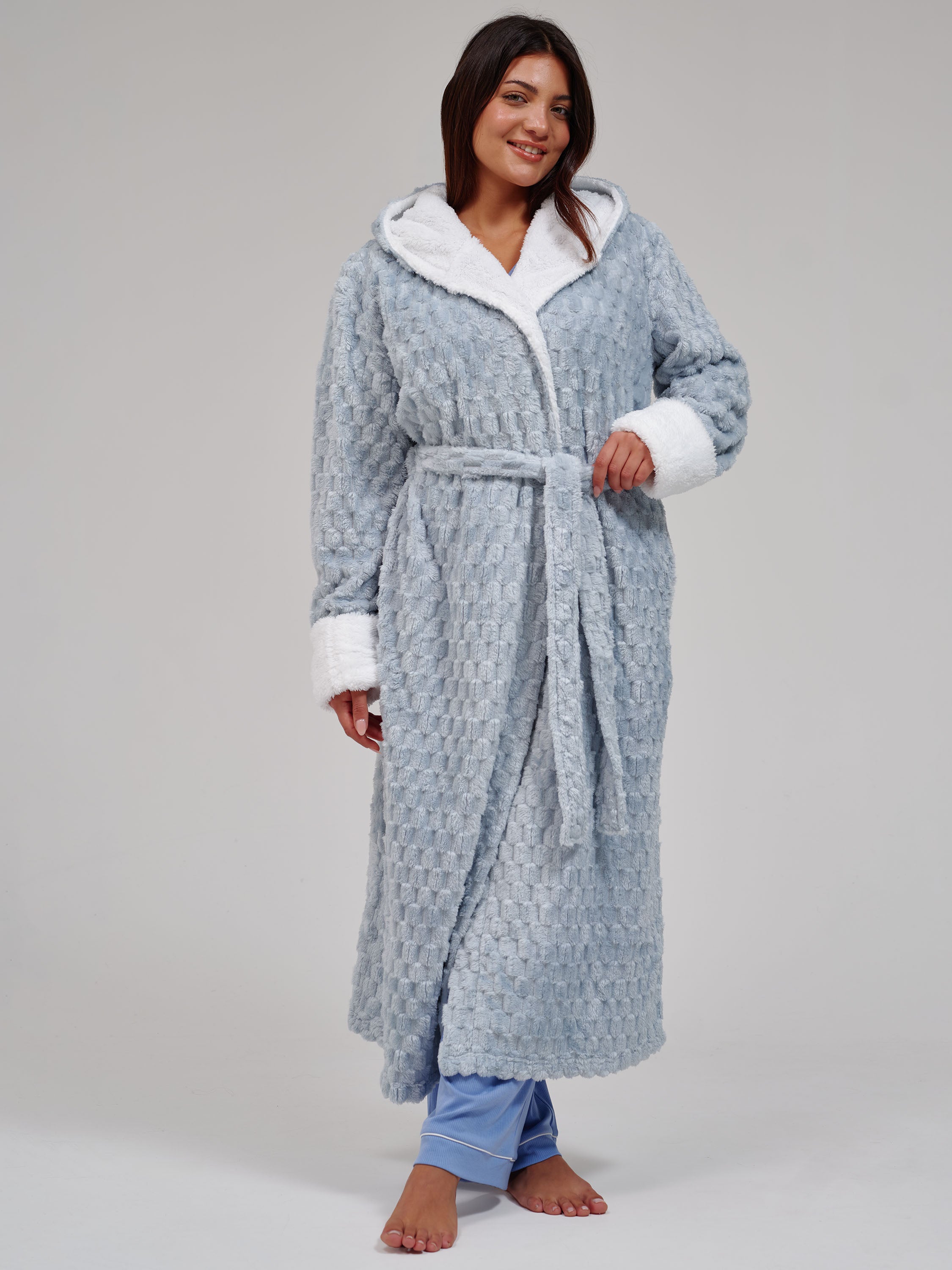 A woman modeling a light blue textured waffle-weave hooded bathrobe with white faux-fur trim on the hood, cuffs, and waist tie, paired with blue pajama pants against a neutral gray studio background.