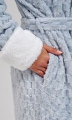 Close-up of a plush light blue bathrobe with a white sherpa-lined cuff and a matching belt, shown on a model with one hand in the robe pocket.