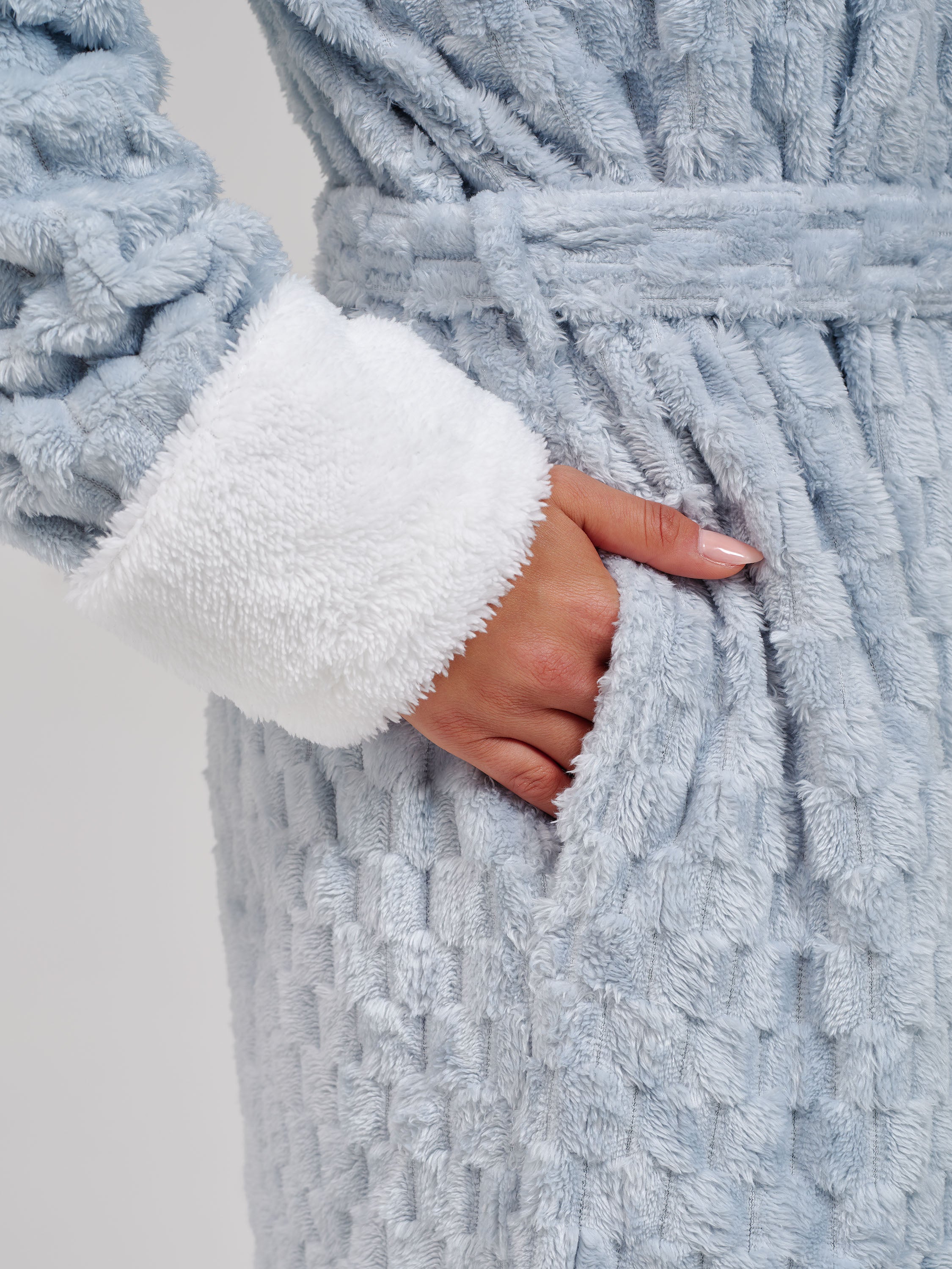 Close-up of a plush light blue bathrobe with a white sherpa-lined cuff and a matching belt, shown on a model with one hand in the robe pocket.