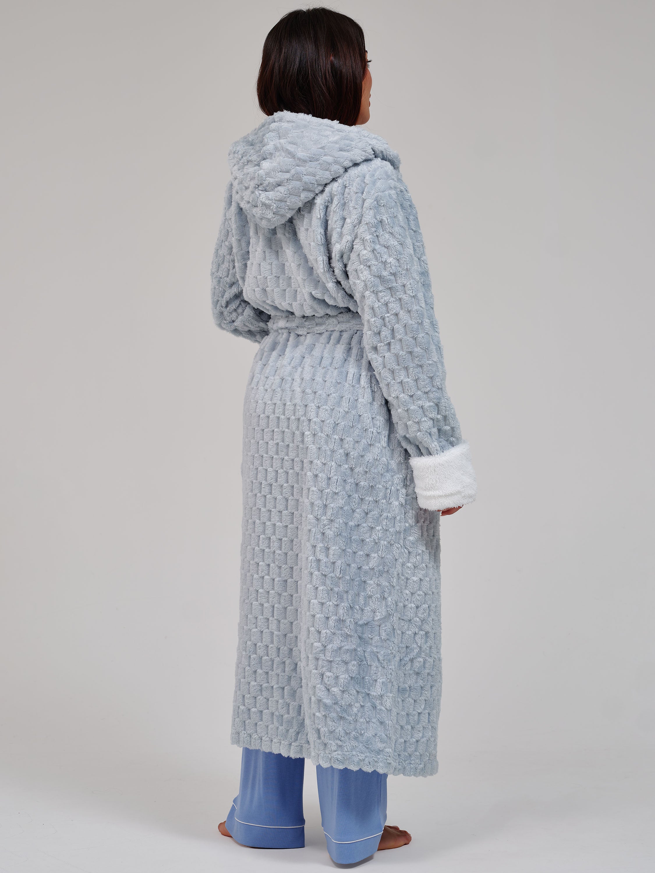 A model shown from the back wearing a light blue textured hooded robe with white cuffs and a matching belt, paired with blue pajama pants, against a neutral gray studio background.