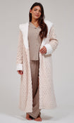 A woman stands barefoot against a light gray studio background wearing a long cream textured robe with white faux-fur trim over a beige pajama set.