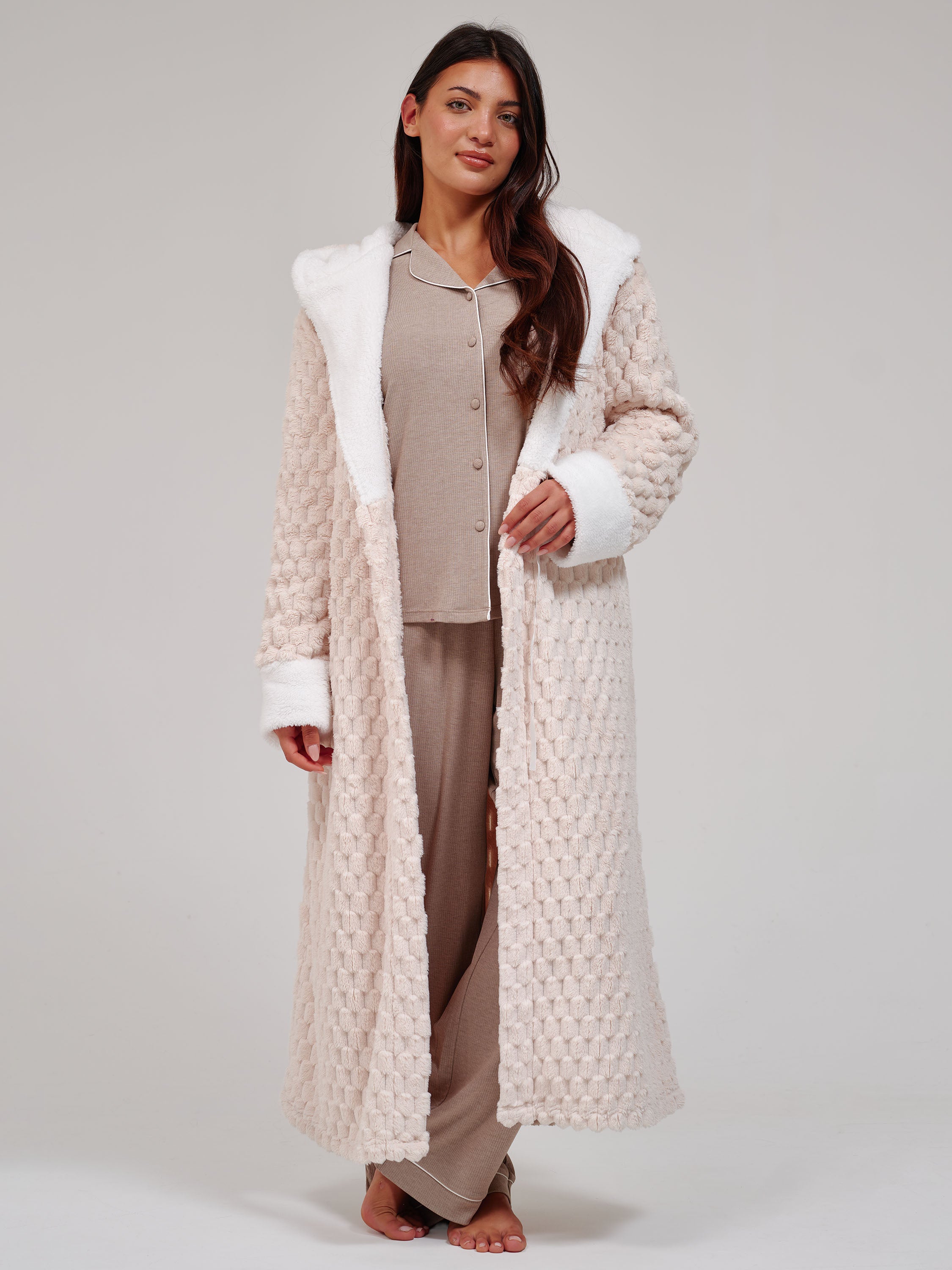 A woman stands barefoot against a light gray studio background wearing a long cream textured robe with white faux-fur trim over a beige pajama set.