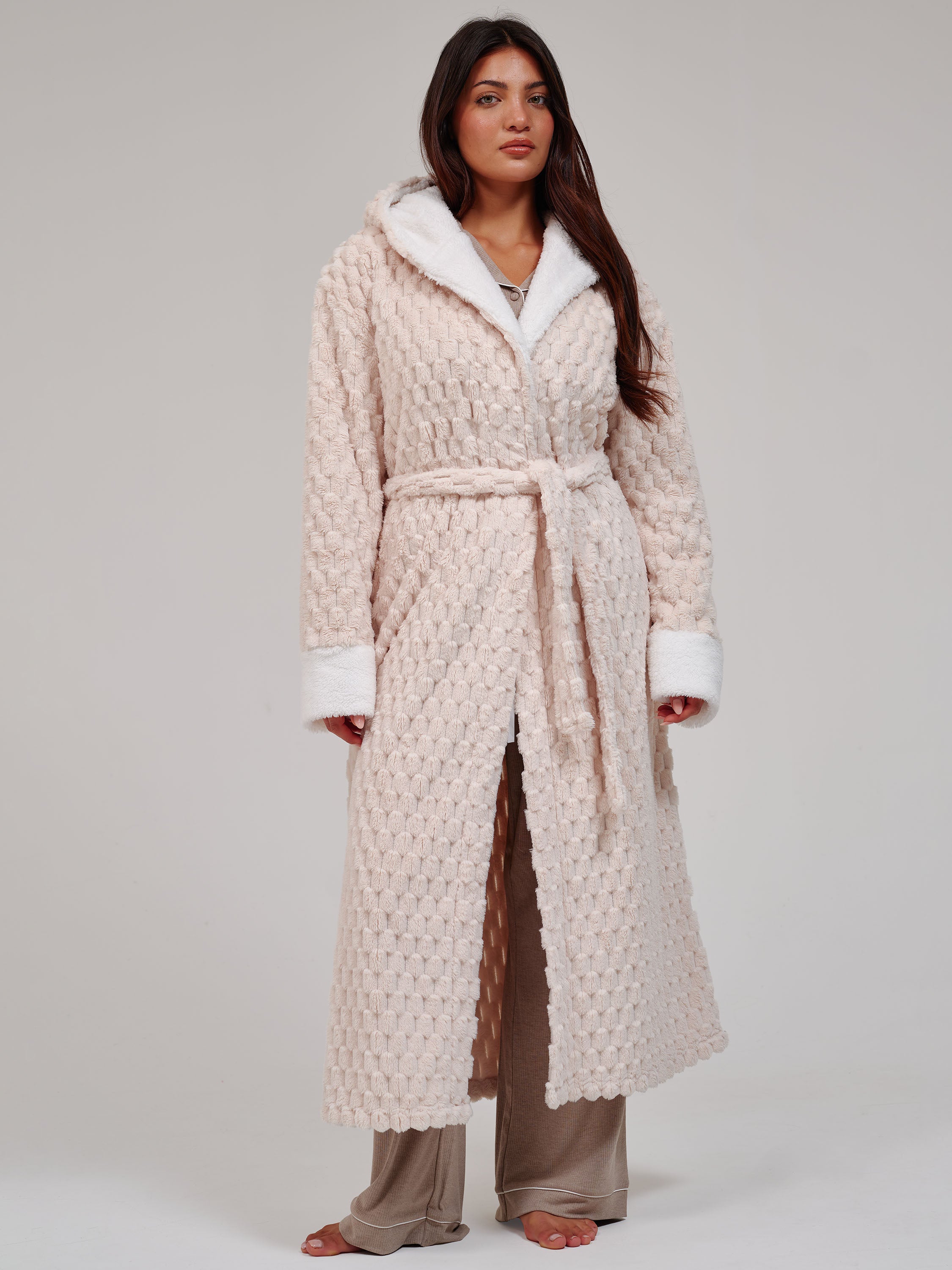 A woman modeling a long cream-colored textured robe with white faux-fur trim, worn over brown lounge pants against a neutral light gray studio background.