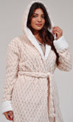 A woman wearing a chunky knit hooded bathrobe in a light cream color with white faux-fur trim, photographed against a neutral light gray studio background.