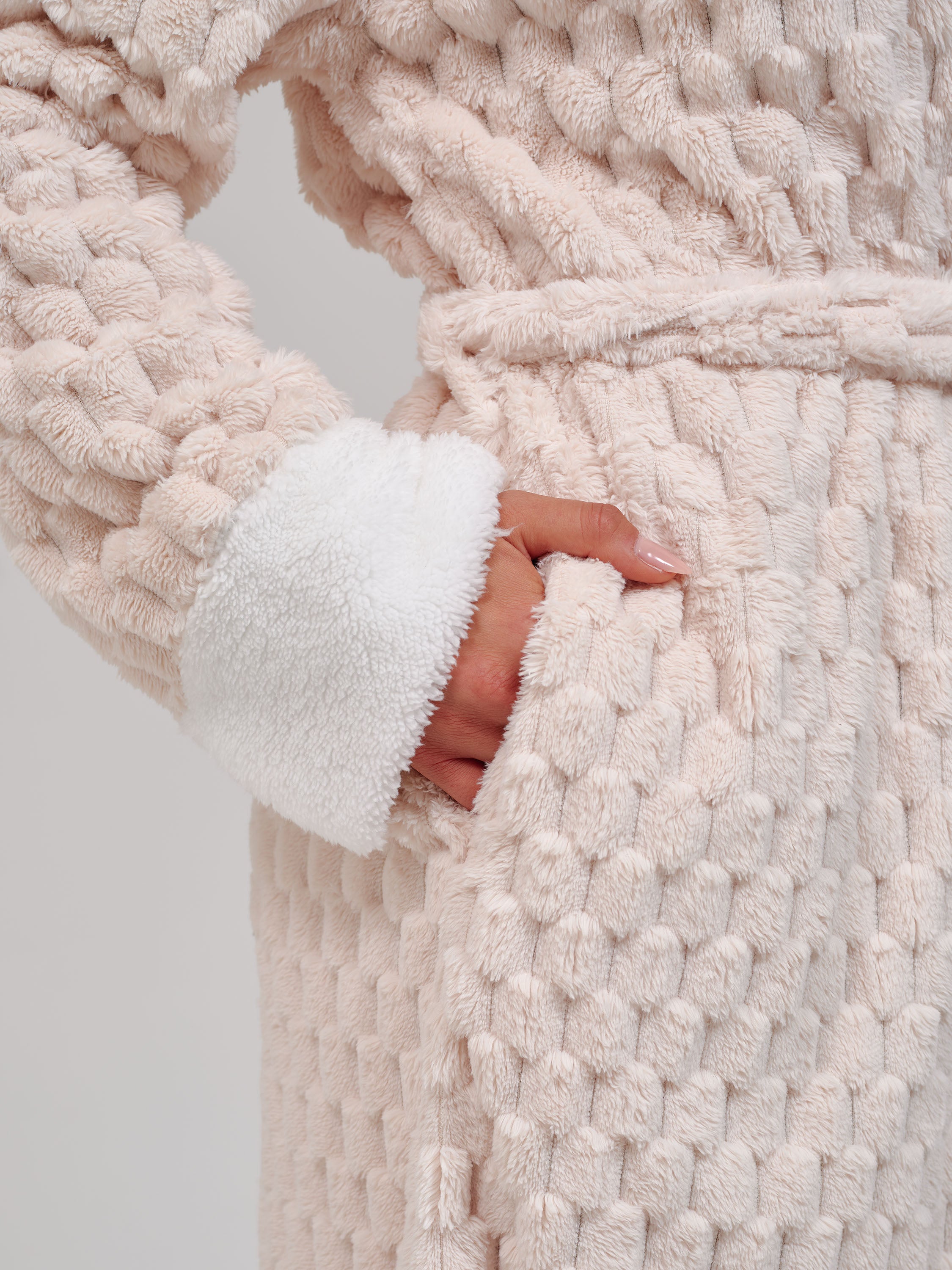 Close-up of a textured cream-colored plush robe with a white sherpa-lined cuff and a hand tucked into a front pocket.