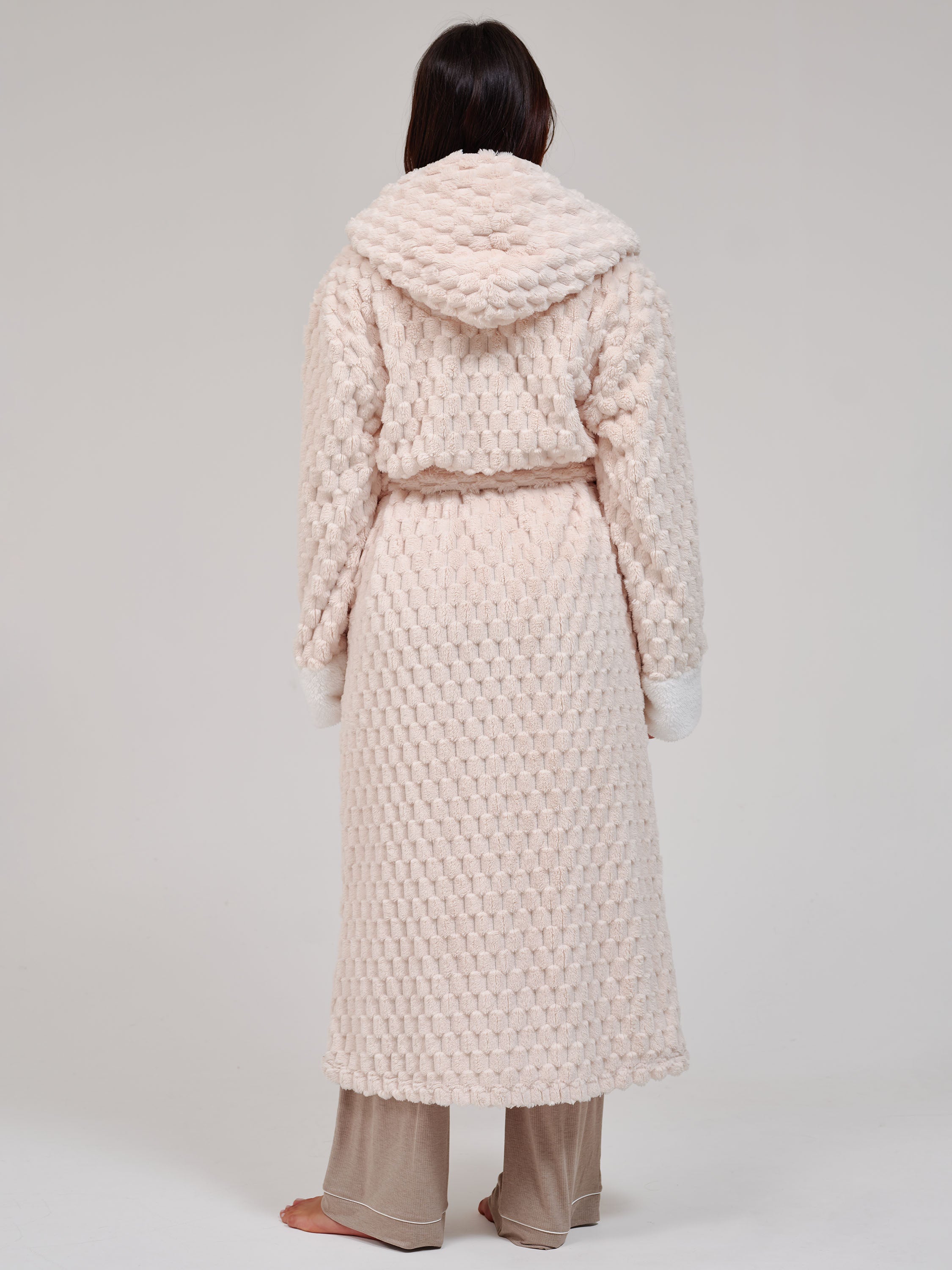 A person shown from the back wearing a long, textured hooded robe in a very light cream tone, paired with beige pants, against a plain light gray studio background.