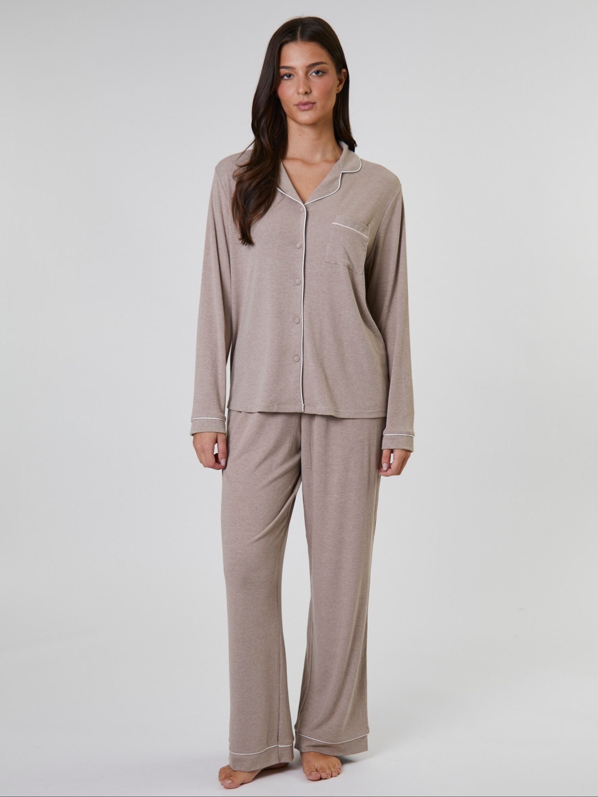 A woman modeling a two-piece pajama set in a light taupe-beige color, featuring a long-sleeve button-front top with a collar and matching wide-leg pants, shot against a plain light gray studio background.
