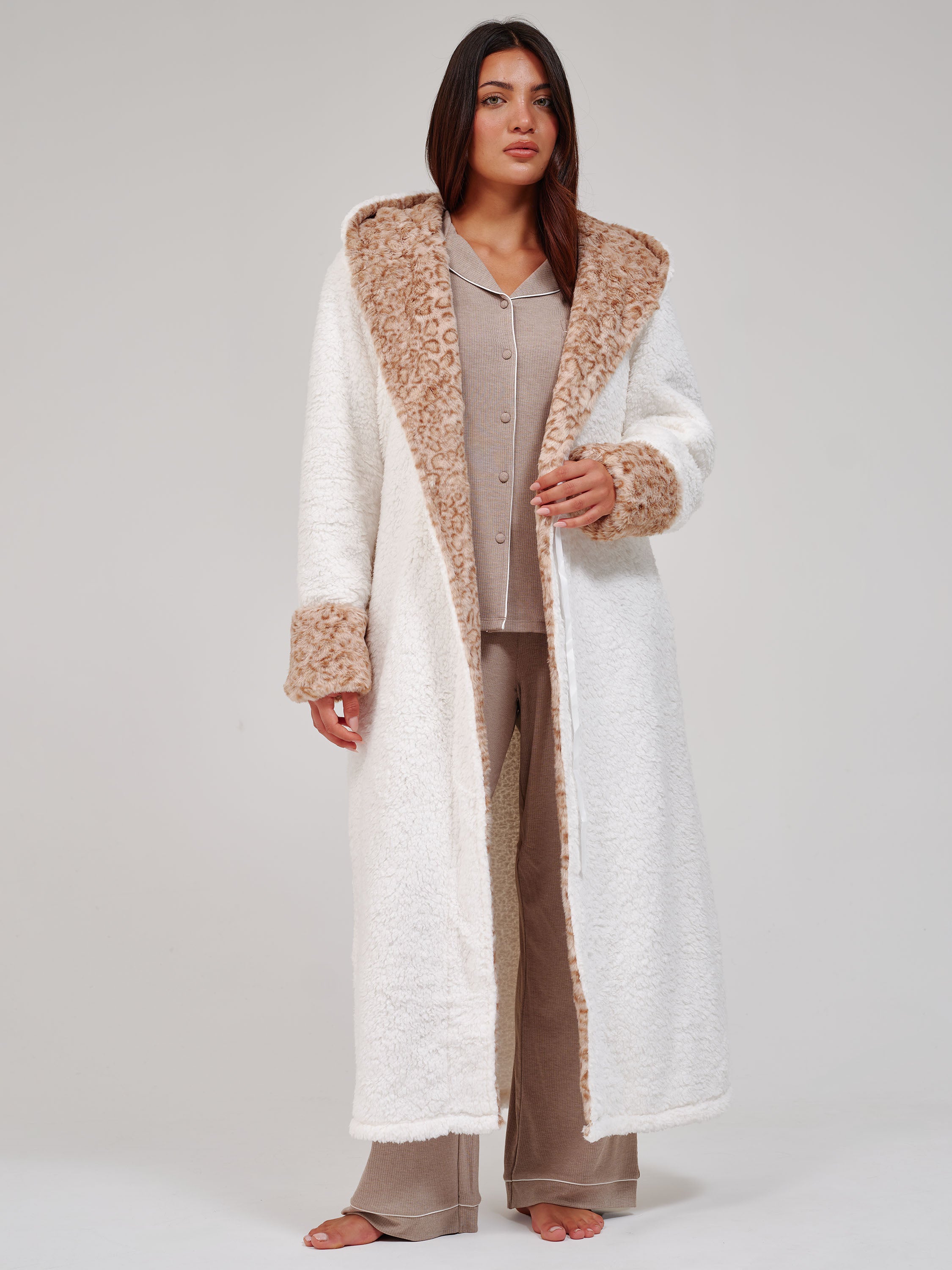 A woman wearing a plush white bathrobe with a leopard patterned hood and cuffs, posed against a neutral light gray background.
