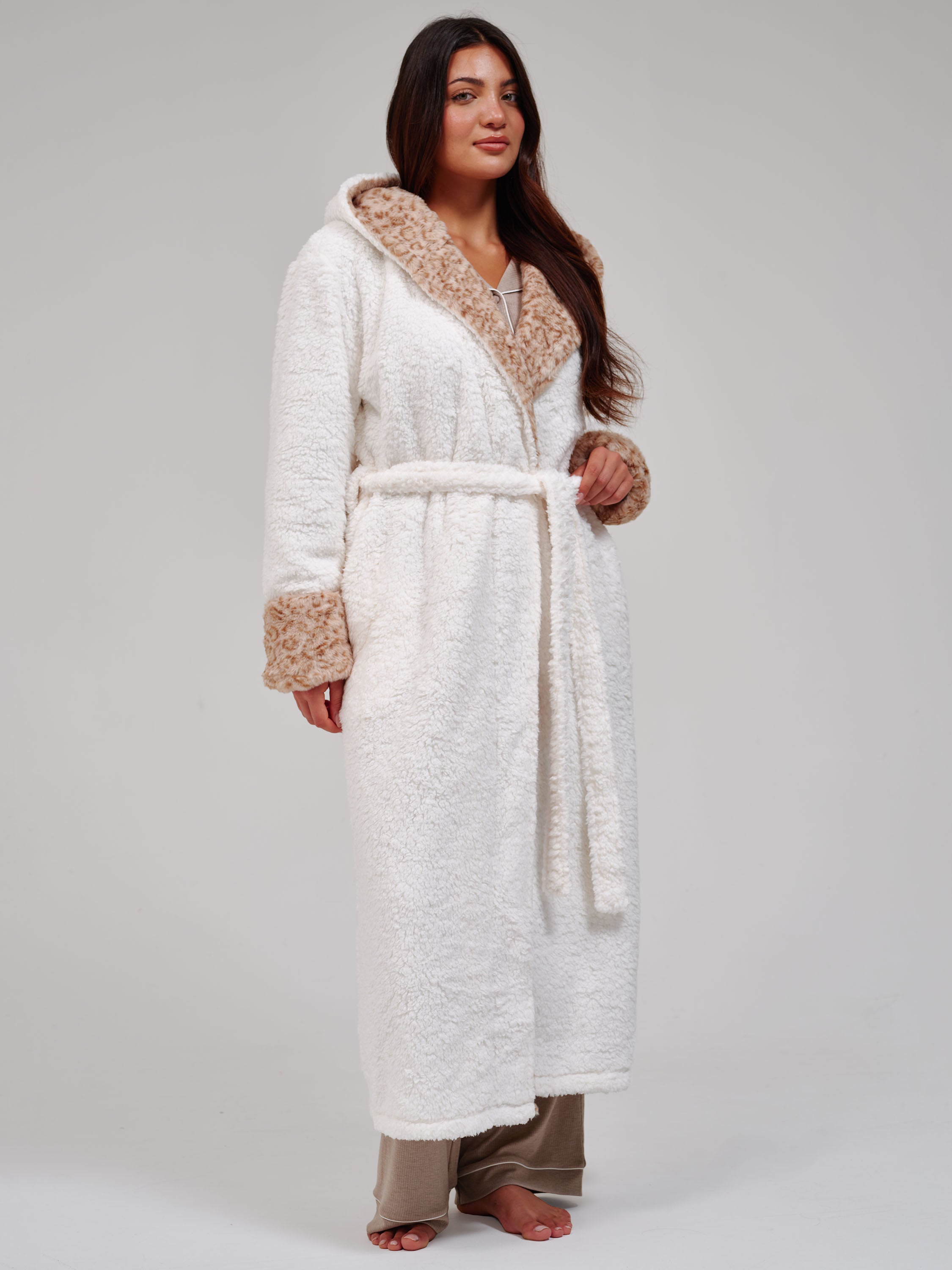A woman wearing a plush white bathrobe with a leopard patterned hood and cuffs, posed against a neutral light gray background.