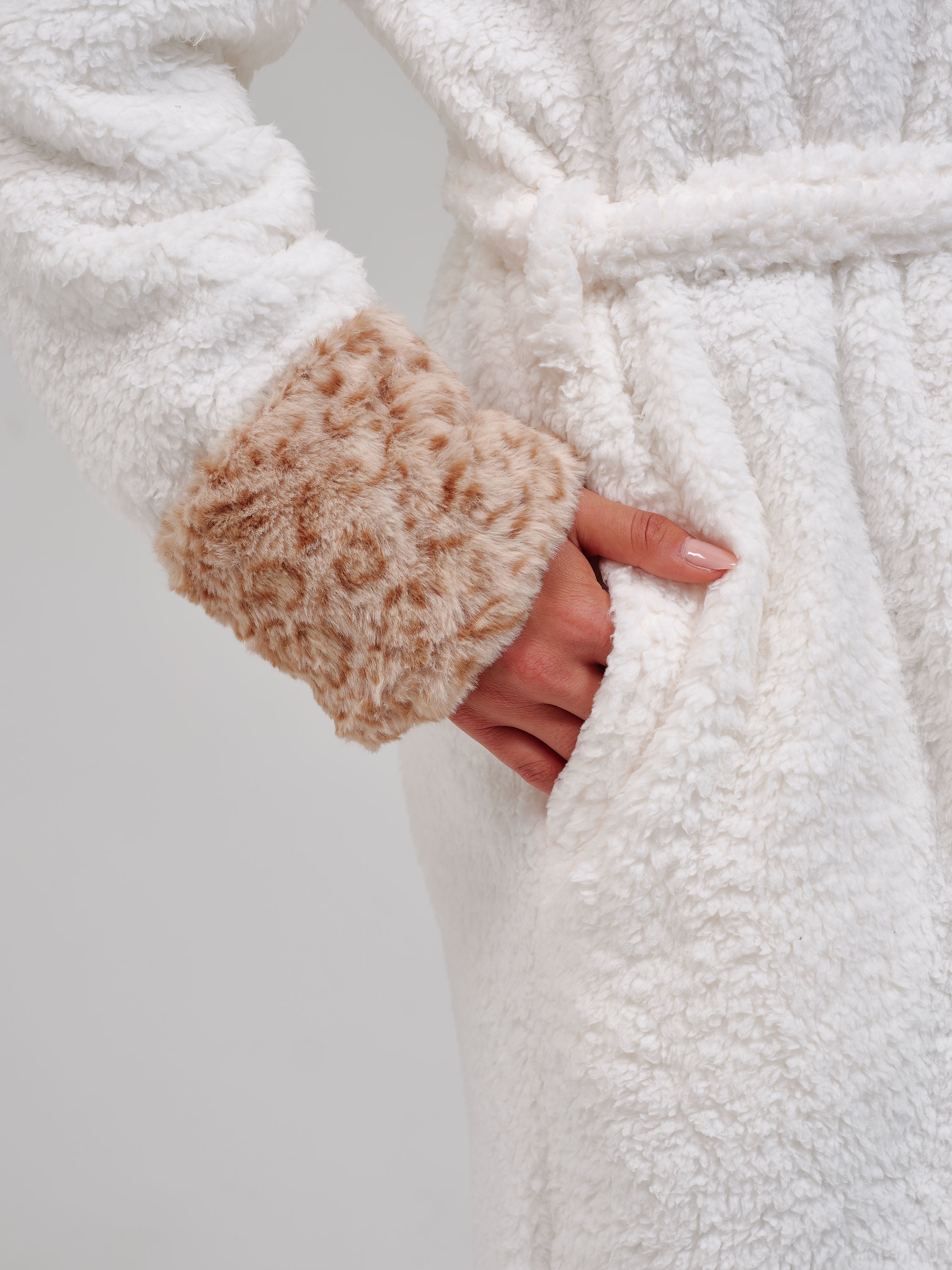 A woman wearing a plush white bathrobe with a leopard patterned hood and cuffs, posed against a neutral light gray background.