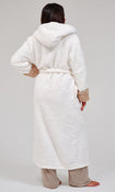 A woman shown from the back wearing a plush white hooded robe with tan textured cuffs and a matching tie belt, paired with beige pants, against a light gray studio background.