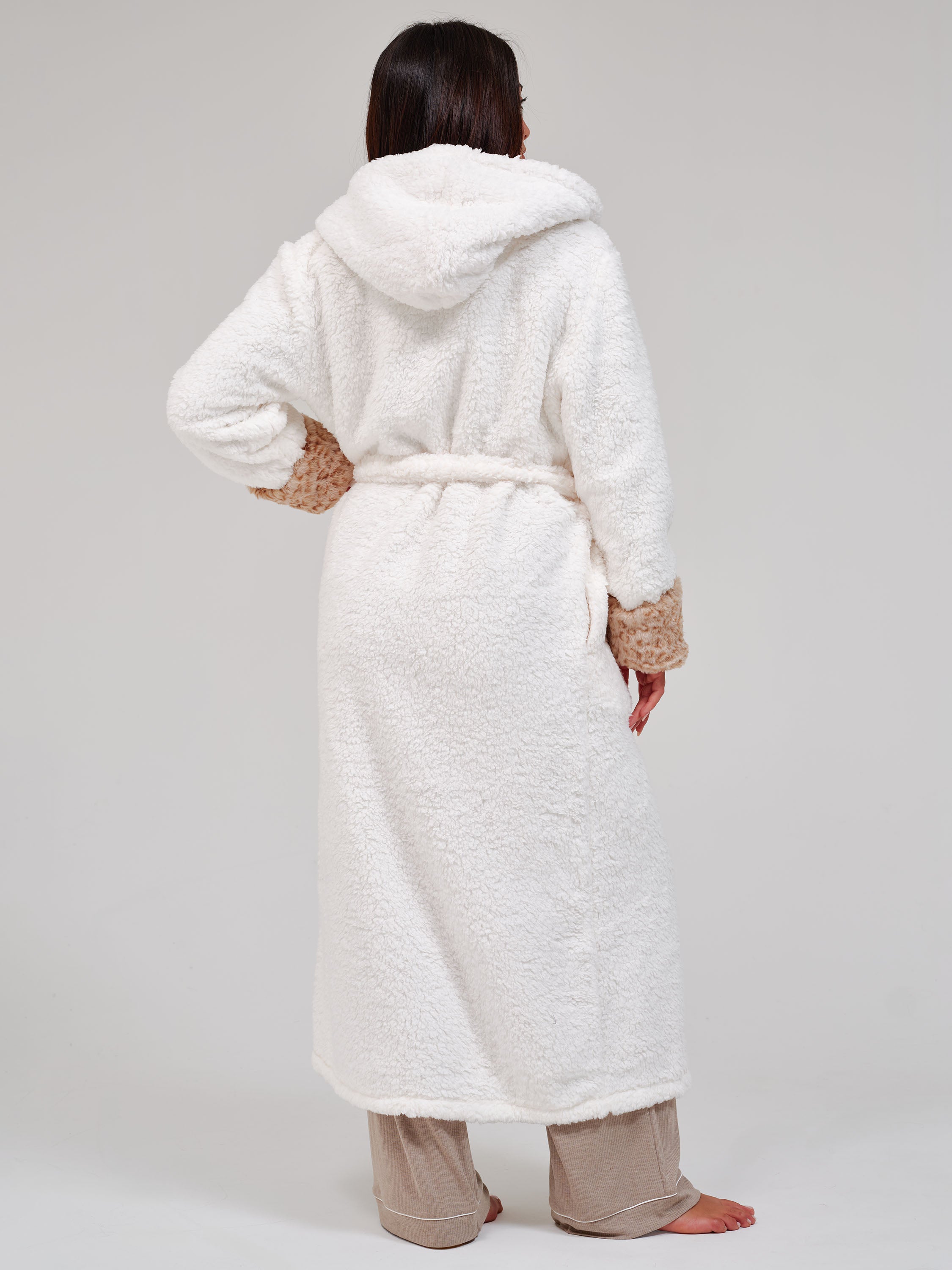 A woman shown from the back wearing a plush white hooded robe with tan textured cuffs and a matching tie belt, paired with beige pants, against a light gray studio background.