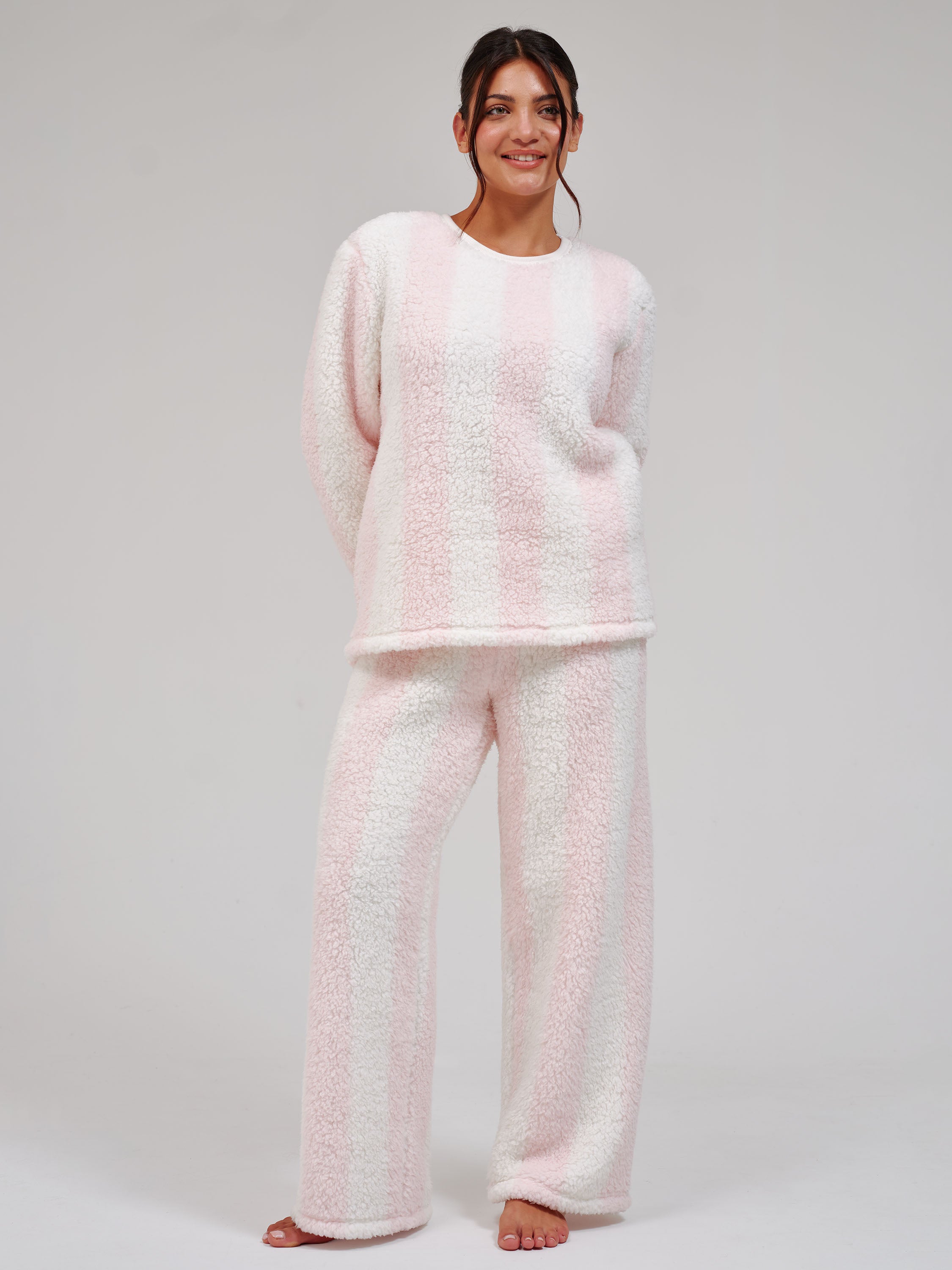 A woman modeling a two-piece fuzzy fleece pajama set with light pink and white vertical stripes, consisting of a long-sleeve top and matching wide-leg pants, against a light gray studio background.