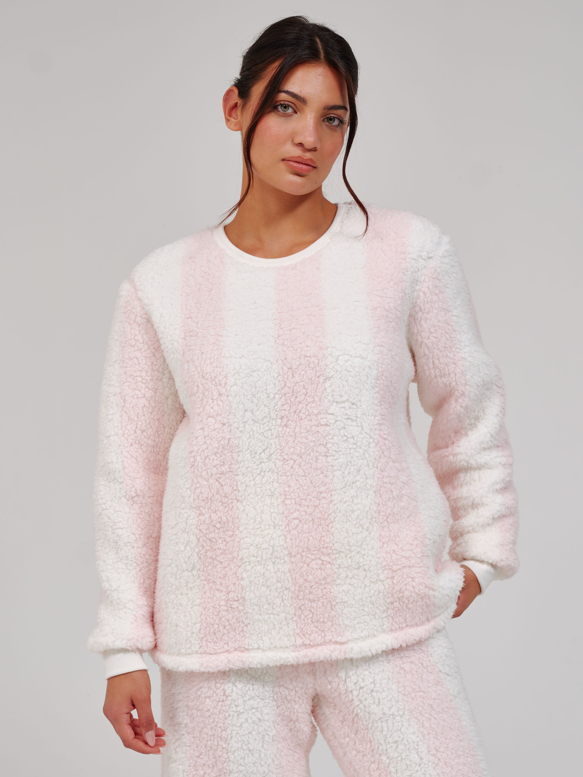 A woman modeling a fuzzy, sherpa-textured long-sleeve sweatshirt and matching sweatpants set in a light pink and white vertical stripe pattern, shot against a solid light gray studio background.
