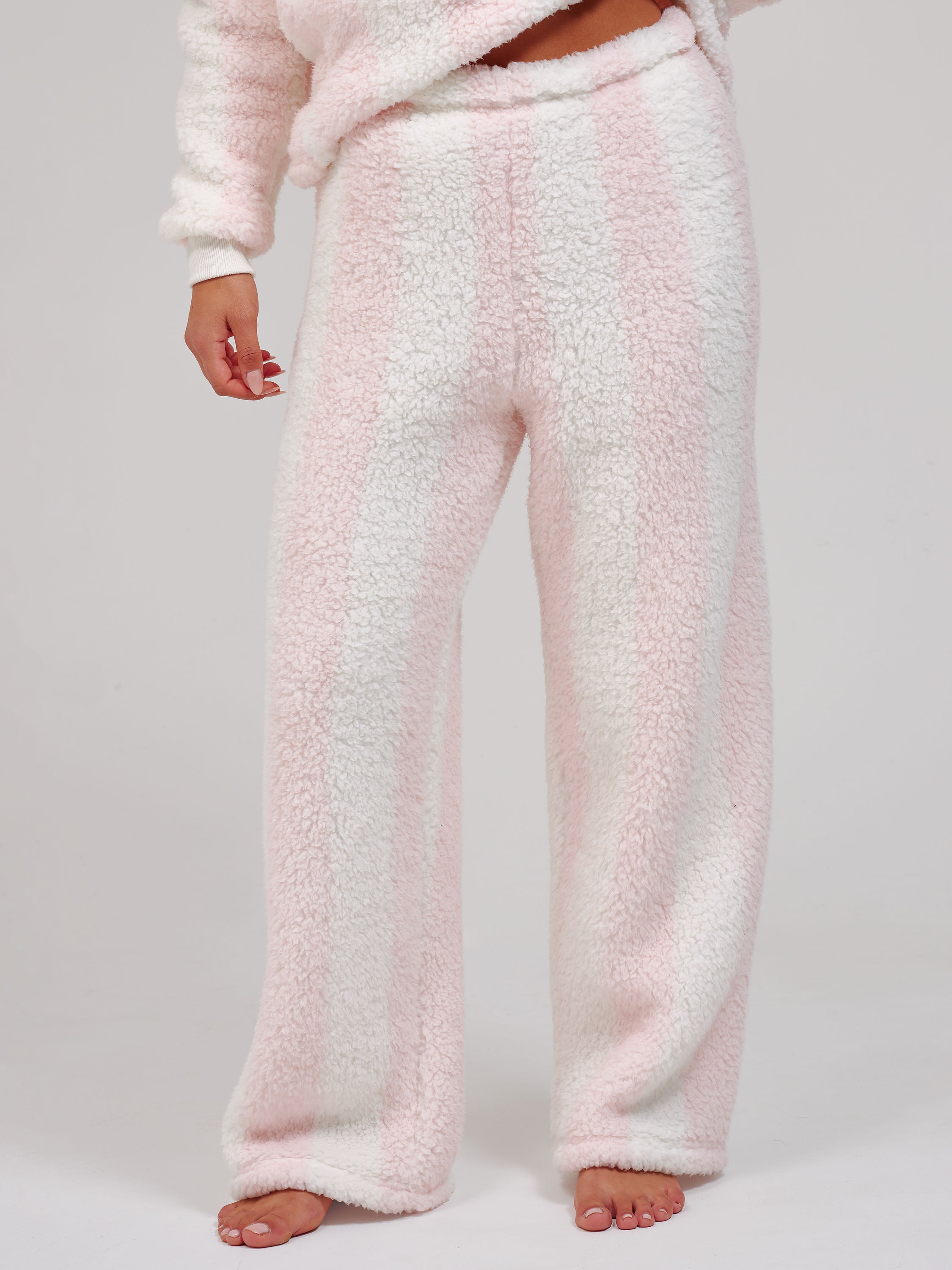 A person wearing plush, wide-leg fleece pajama pants in a pale pink and white striped pattern, shown from the waist down against a light gray studio background.