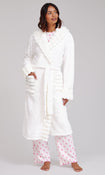 A woman modelling a plush white bathrobe with a shawl collar and matching white pyjama pants with a pink cowboy boot pattern, standing against a light grey studio background.