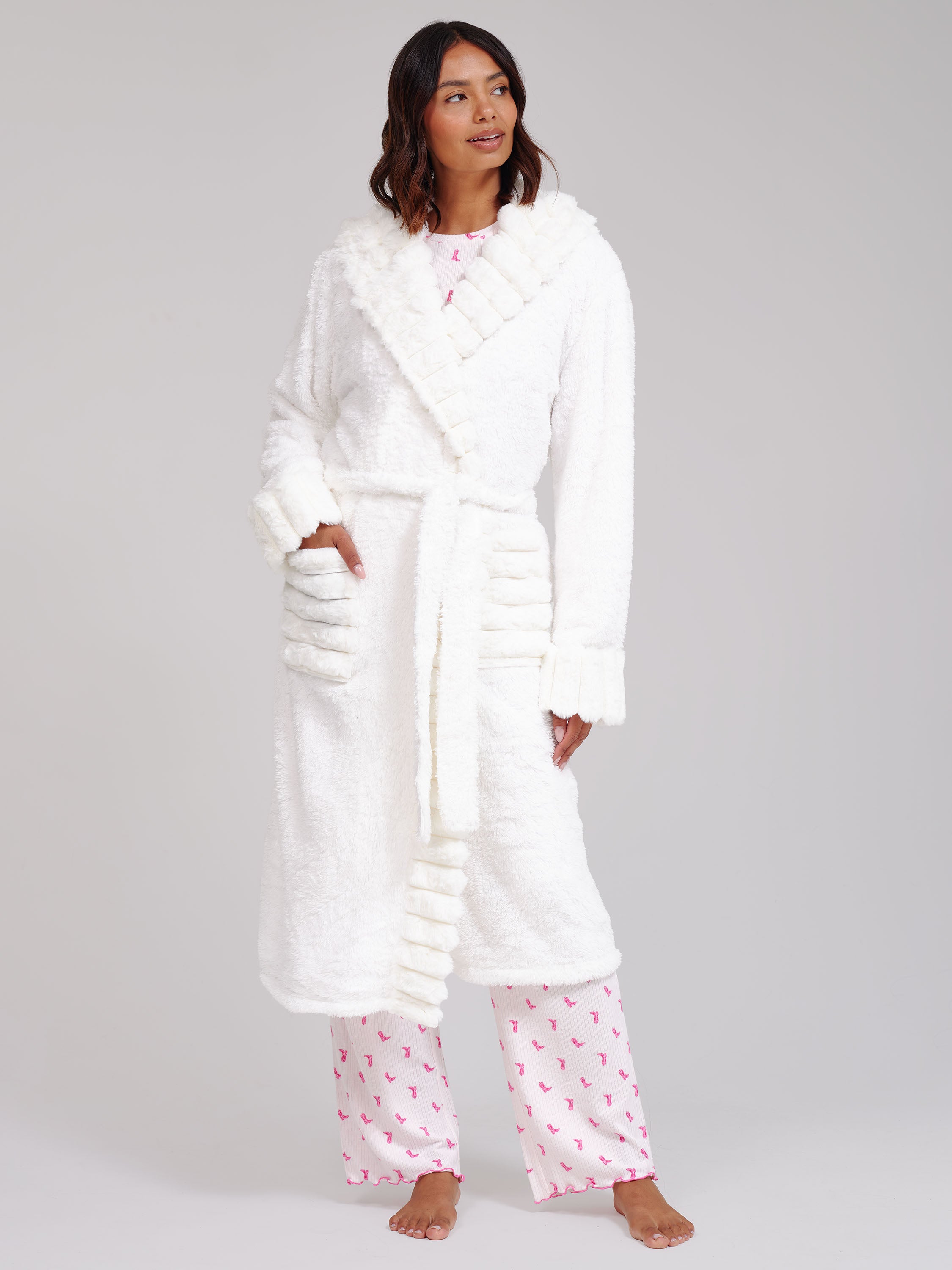 A woman modelling a plush white bathrobe with a shawl collar and matching white pyjama pants with a pink cowboy boot pattern, standing against a light grey studio background.
