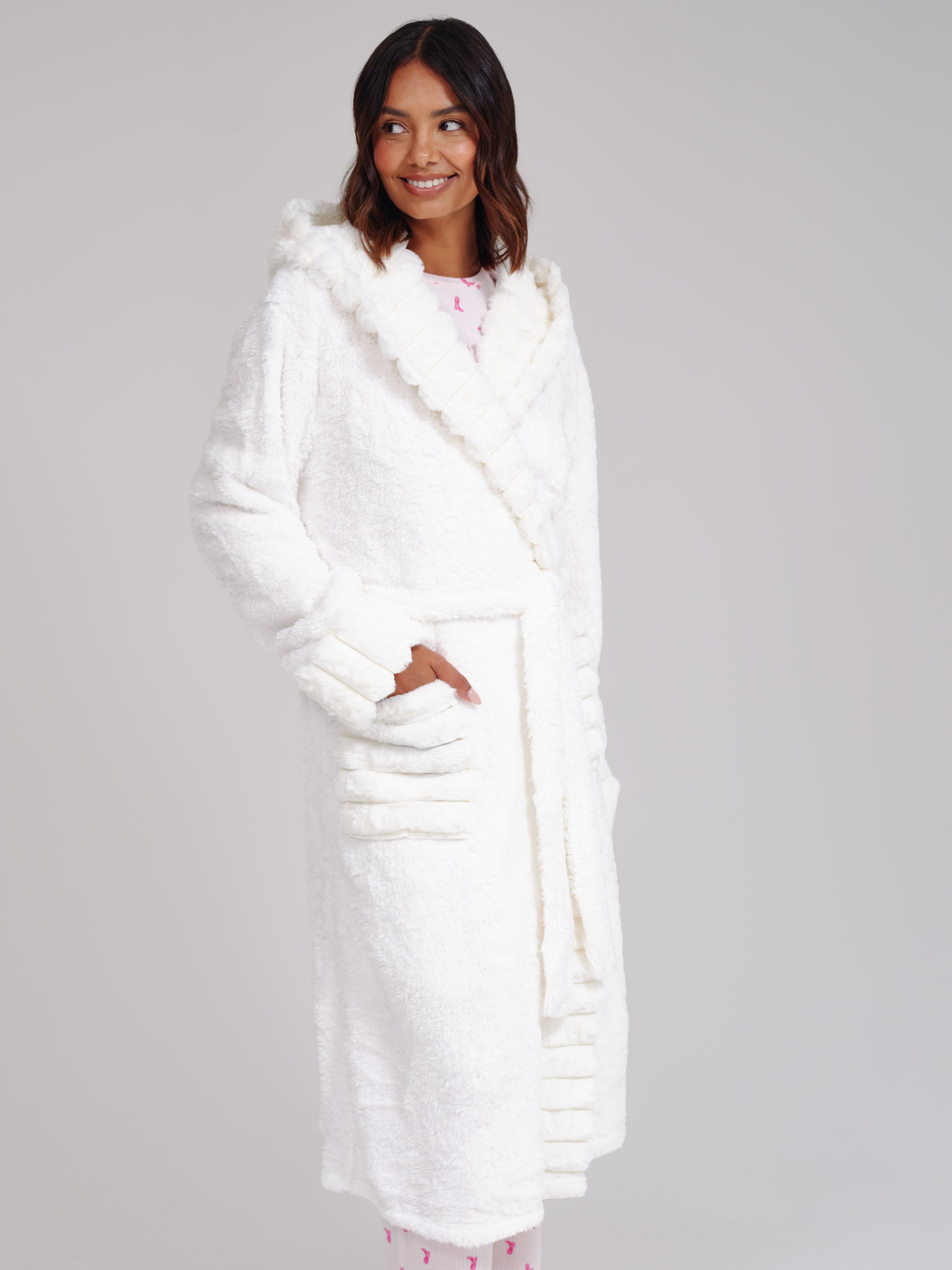 A woman wearing a plush white hooded bathrobe with faux-fur trim and front pockets, standing against a light gray studio background.