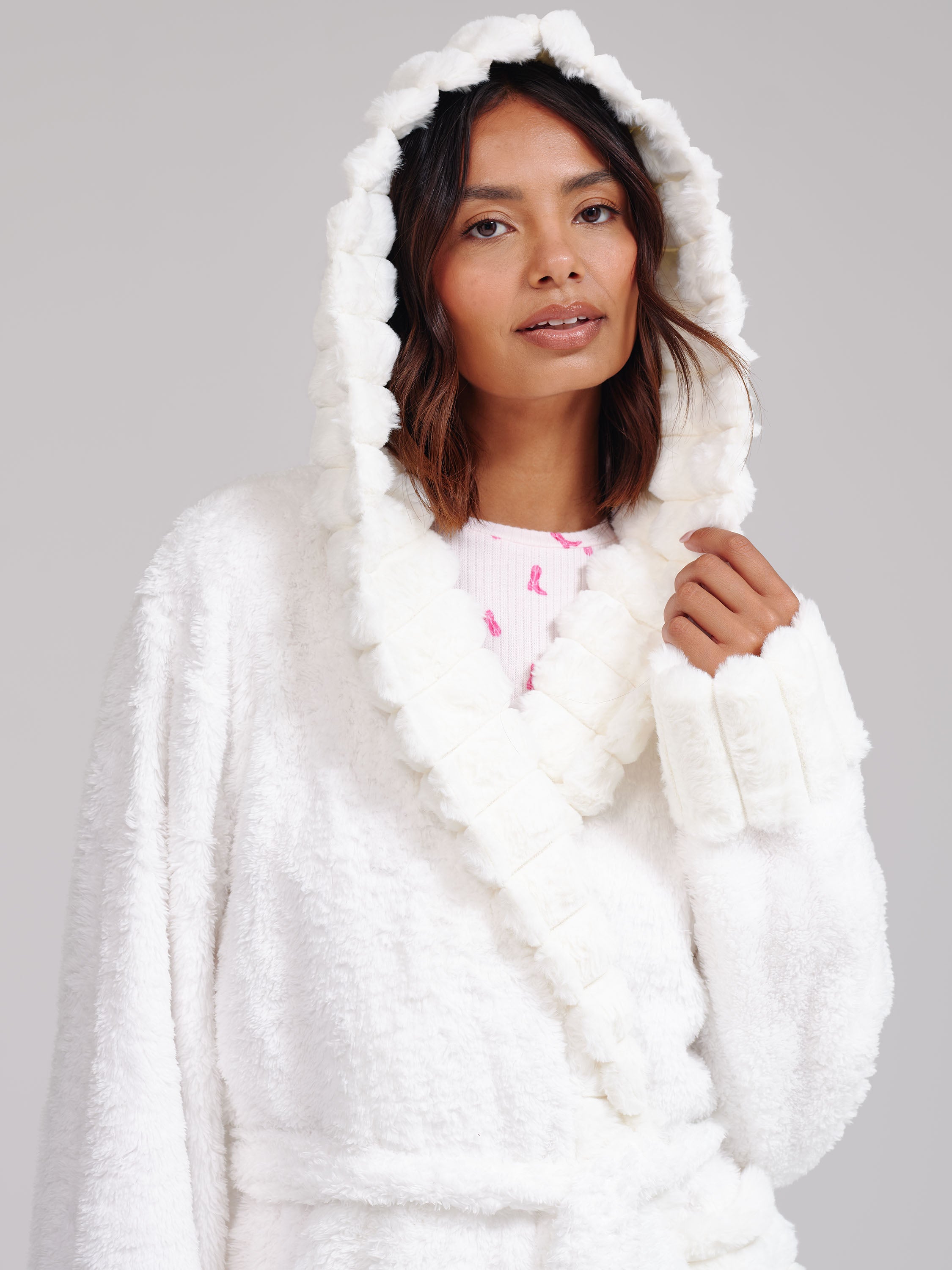 A woman wearing a plush white hooded robe with a shawl collar, photographed against a plain light gray studio background.