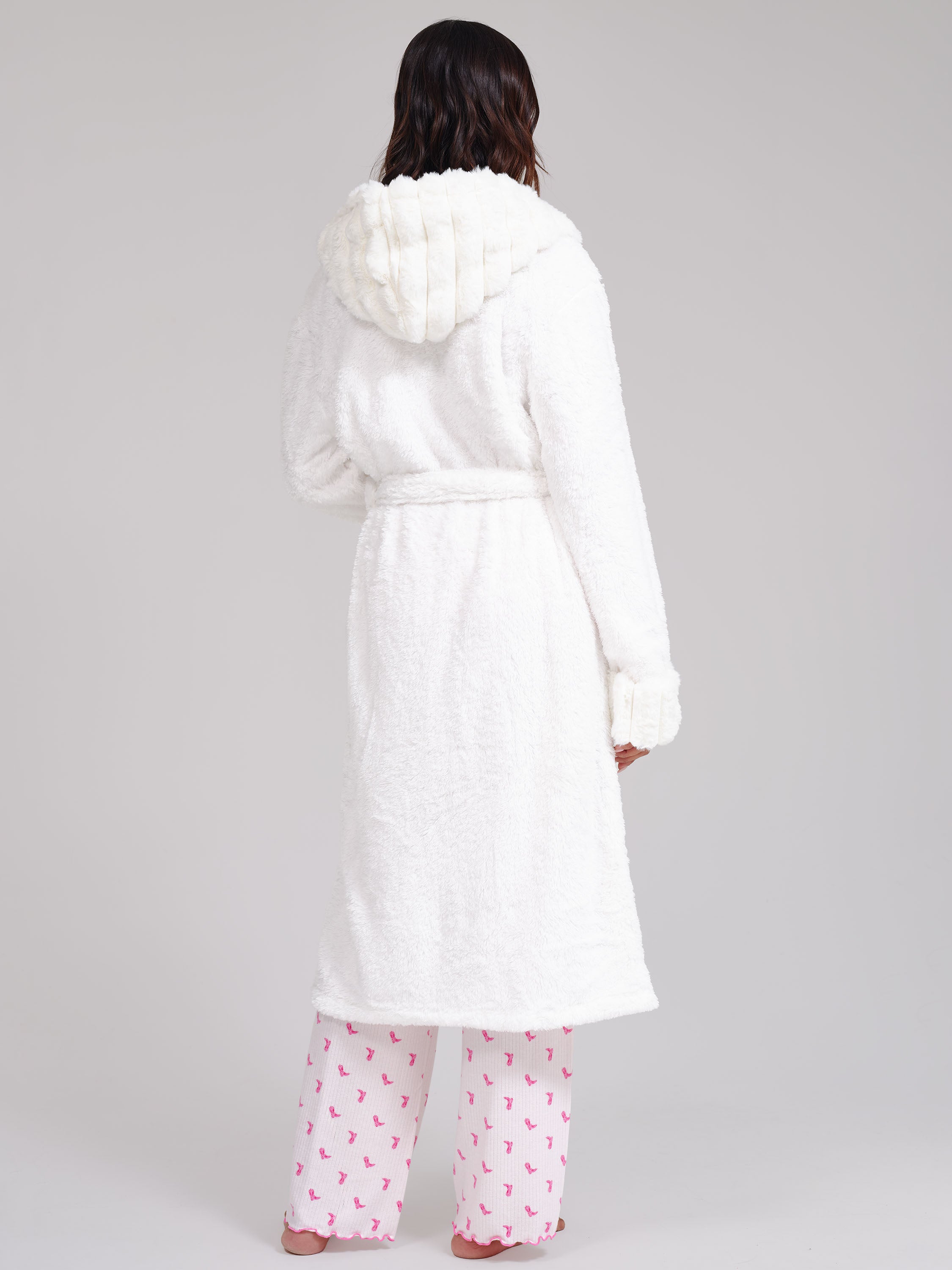 A woman shown from the back wearing a plush white hooded robe with a tie belt, paired with pink patterned pajama pants, against a light gray studio background.