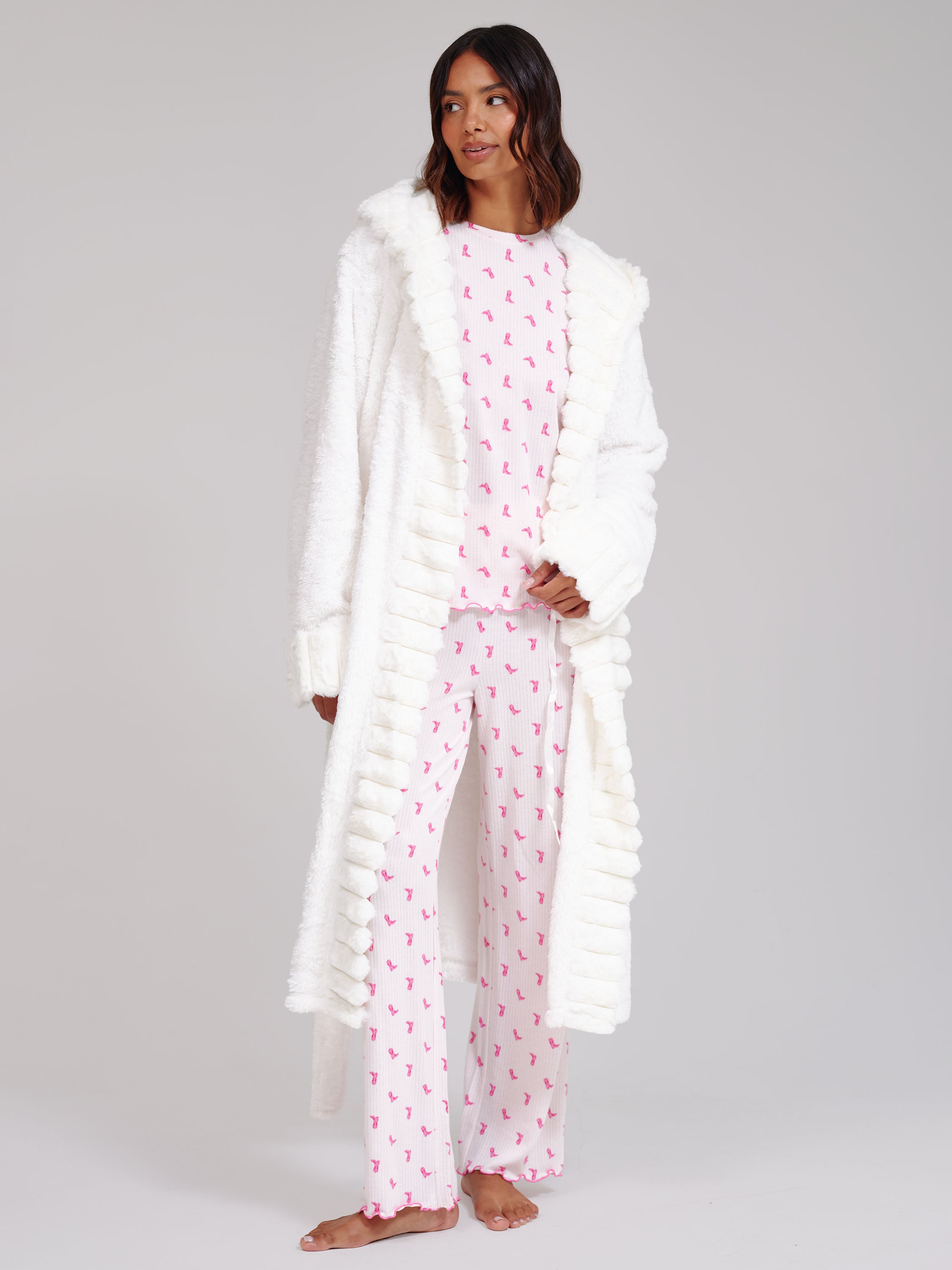 A woman modelling a two-piece pajama set with a long white faux-fur robe. The pajama set consists of a light pink long-sleeve top and matching pants, both patterned with small pink cowboy boot motifs.
